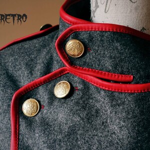 May include: Close-up of a gray wool jacket with a red leather trim and gold buttons. The jacket has a double-breasted closure and a high collar.