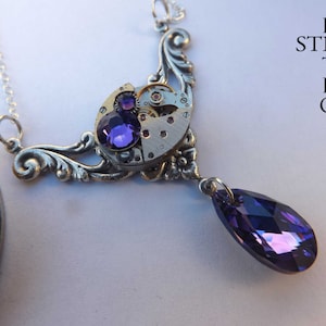 May include: A silver necklace with a steampunk-style pendant featuring a watch face with purple rhinestones and a dangling purple crystal.