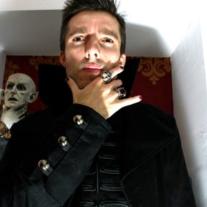 May include: A man wearing a black coat with silver buttons and a black shirt with a decorative design. He is wearing several rings on his fingers. The text "STEAMRETRO" is visible in the background.