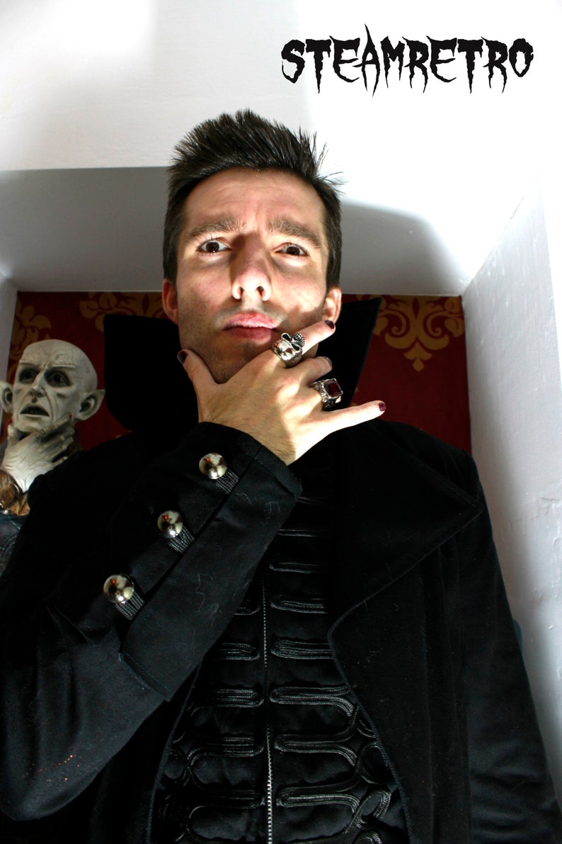 May include: A man wearing a black coat with silver buttons and a black shirt with a decorative design. He is wearing several rings on his fingers. The text "STEAMRETRO" is visible in the background.