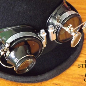 Peut inclure: Lunettes steampunk noires avec des lentilles grossissantes et un cadre en m&eacute;tal argent&eacute;. Les lunettes sont attach&eacute;es &agrave; un chapeau noir.