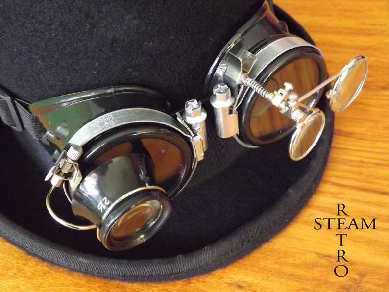 Peut inclure: Lunettes steampunk noires avec des lentilles grossissantes et un cadre en m&eacute;tal argent&eacute;. Les lunettes sont attach&eacute;es &agrave; un chapeau noir.
