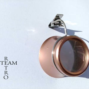 Puede incluir: Una lupa de oro rosa con un clip en la parte superior. La lupa está unida a un marco de metal.