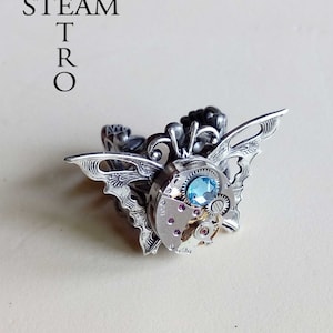 Könnte beinhalten: Ein silberner Ring mit einem Schmetterlingsdesign, das mit einem blauen Edelstein und komplizierten Uhrwerkdetails verziert ist.