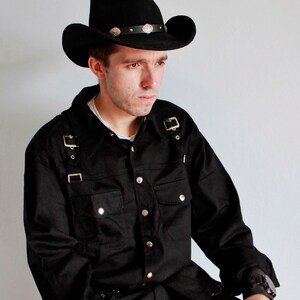 May include: A person wearing a black cowboy hat with silver accents, a black button-up shirt with multiple straps, and black pants. The shirt has a pocket on the left side and the pants have a belt with metal buckles. The person is sitting down and looking to the side.