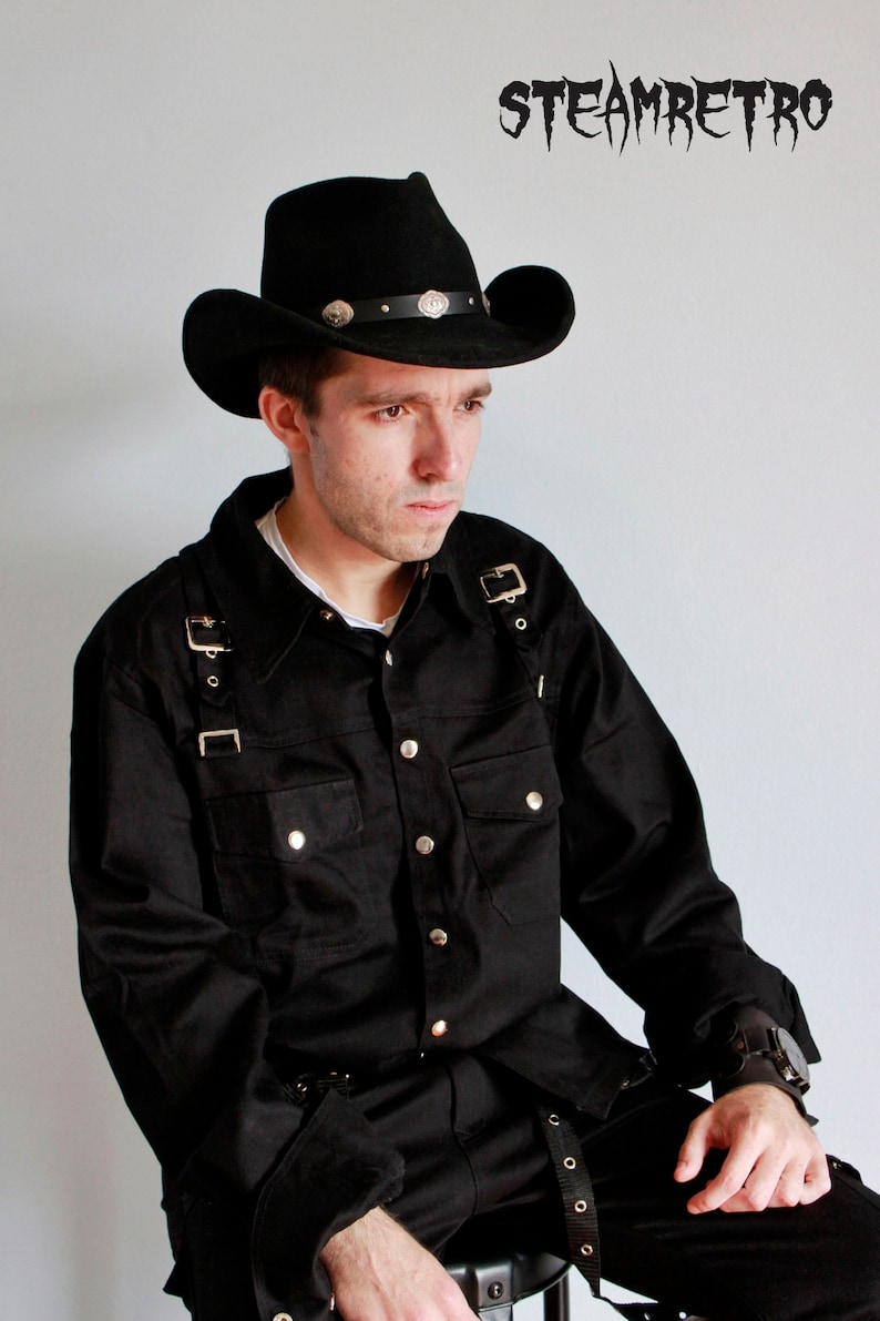 May include: A person wearing a black cowboy hat with silver accents, a black button-up shirt with multiple straps, and black pants. The shirt has a pocket on the left side and the pants have a belt with metal buckles. The person is sitting down and looking to the side.