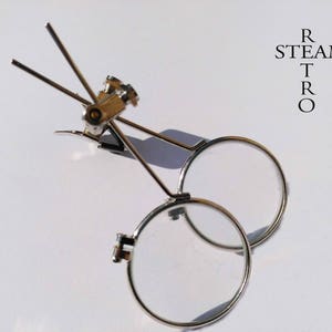 Könnte beinhalten: Ein Paar silberfarbene Lupen mit runden Gläsern, die an einem Metallrahmen befestigt sind. Der Rahmen hat zwei verstellbare Arme, die sich verlängern lassen, um über den Kopf zu passen.