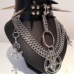 May include: A black leather choker with silver spikes and studs, paired with layered silver chain necklaces. The necklaces feature a large pentagram pendant with a goat's head and other gothic-style charms. The jewelry is displayed on a black mannequin.