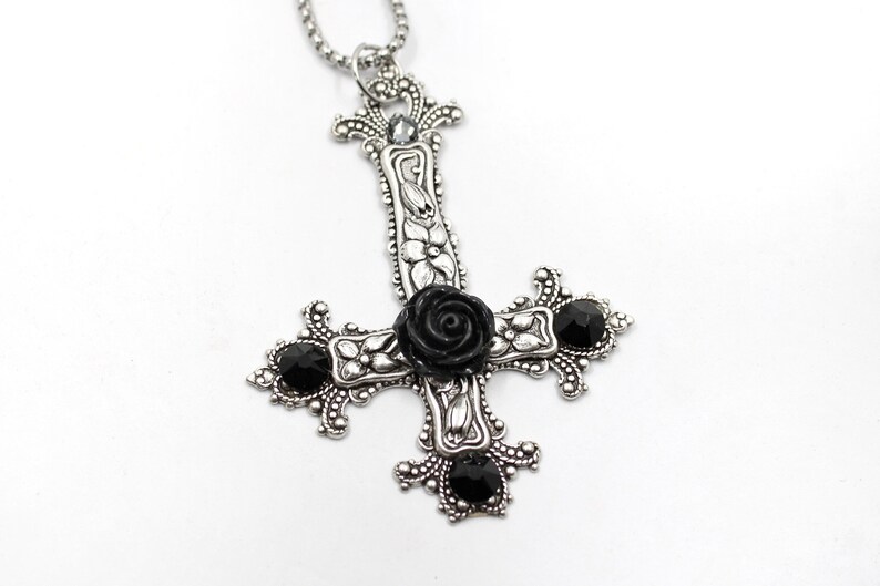 Puede incluir: Un collar con un colgante de plata en forma de cruz invertida adornado con una rosa negra y piedras preciosas negras.