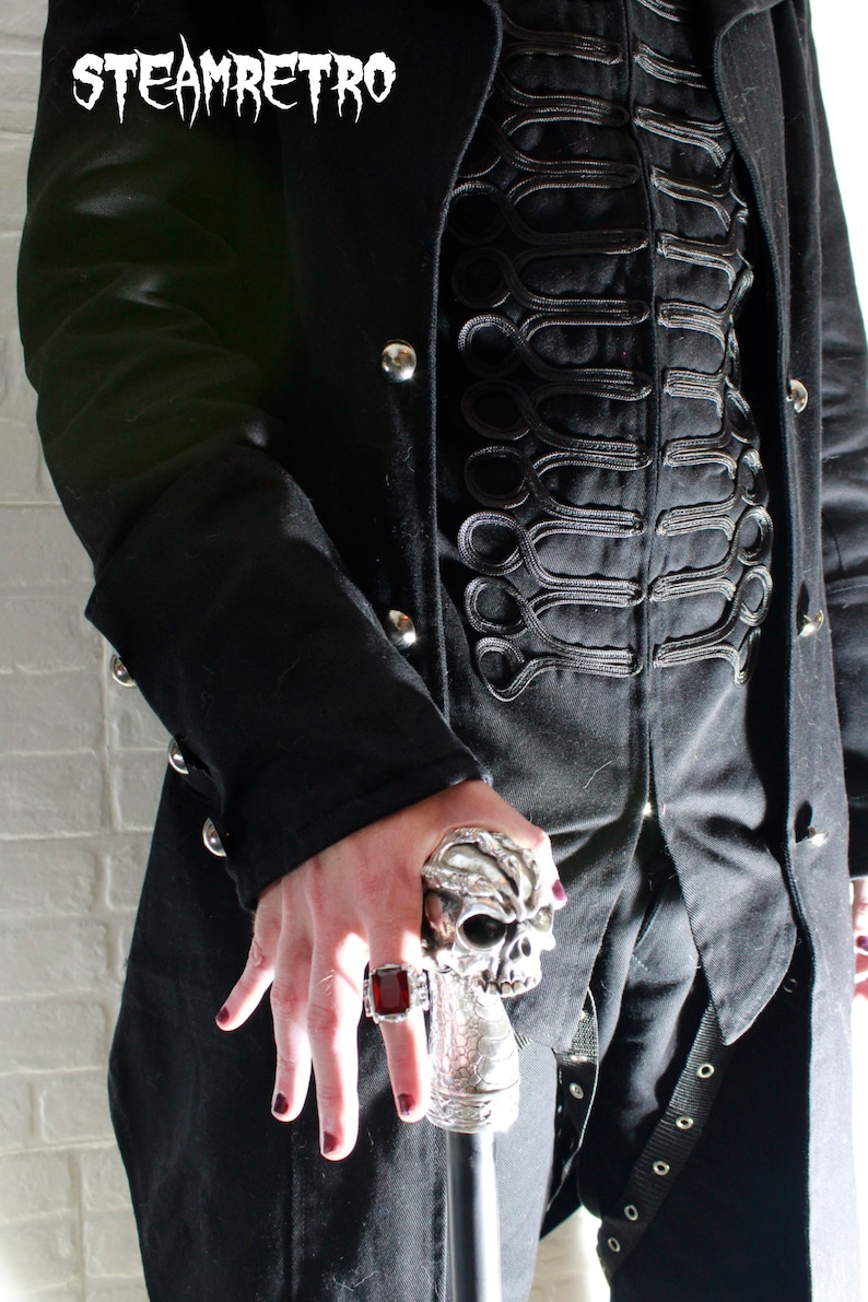May include: A person wearing a black coat with a skull-shaped walking stick. The coat has a decorative trim of black braid. The person is wearing a ring with a red stone.
