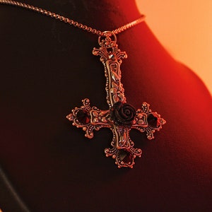 Puede incluir: Un collar con un colgante de plata con una rosa negra y un dise&ntilde;o de pentagrama. El collar est&aacute; en un expositor de terciopelo negro.