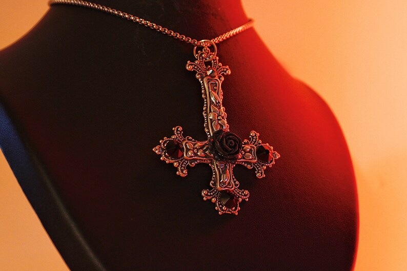 Puede incluir: Un collar con un colgante de plata con una rosa negra y un dise&ntilde;o de pentagrama. El collar est&aacute; en un expositor de terciopelo negro.