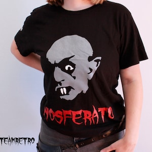 May include: A black t-shirt featuring a white and grey graphic of a vampire's face and the text "Nosferatu" in red.