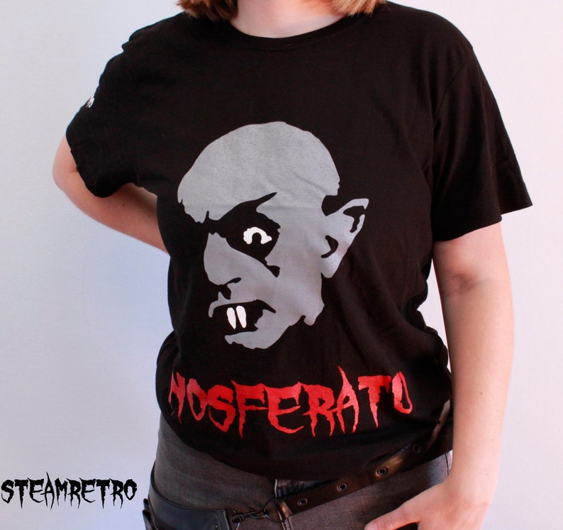 May include: A black t-shirt featuring a white and grey graphic of a vampire's face and the text "Nosferatu" in red.
