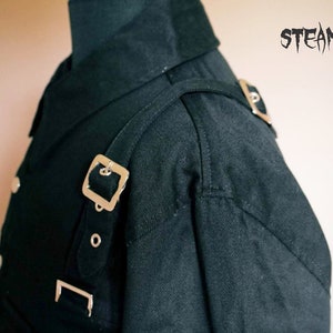 May include: A black collared shirt with a silver buckle strap across the shoulder. The shirt has a double breasted closure and a collar that stands up.