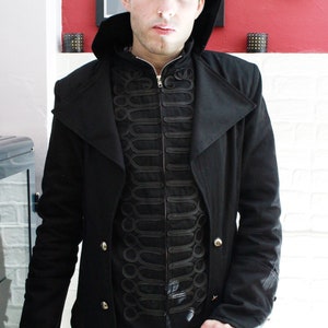 May include: A black long coat with a high collar and a black zippered vest underneath. The vest has decorative black stitching and a gold button. The person is holding a silver skull-shaped walking stick.