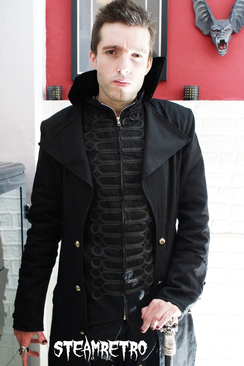 May include: A black long coat with a high collar and a black zippered vest underneath. The vest has decorative black stitching and a gold button. The person is holding a silver skull-shaped walking stick.