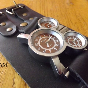 Puede incluir: Un reloj de pulsera de estilo steampunk plateado con tres esferas redondas. La esfera principal tiene un fondo marrón con números y manecillas blancas. El reloj está en una correa de cuero negro con dos hebillas.