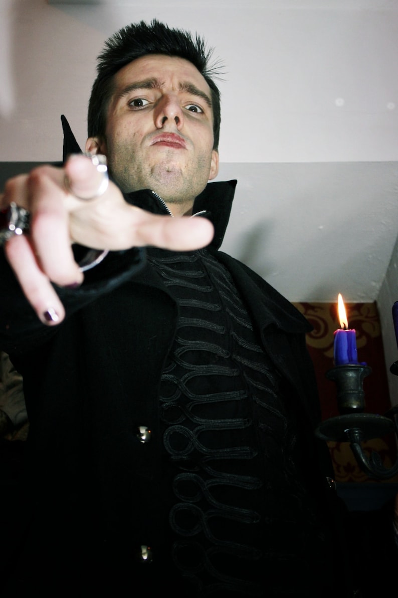 May include: A man in a black coat with a high collar and intricate embroidery. He is pointing at the camera with a serious expression. A purple candle is lit in the background.