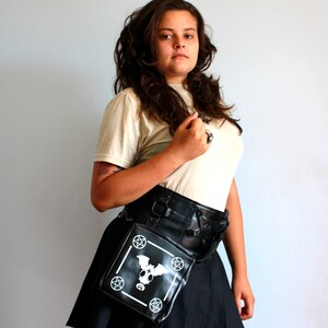 May include: A woman wearing a white t-shirt and a black skirt with a black leather belt and a black bag with a white bat and pentagram design. The bag is worn on the hip.