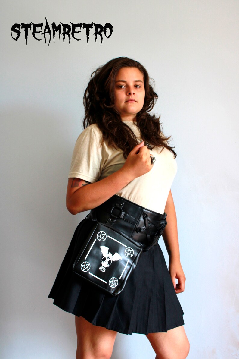 May include: A woman wearing a white t-shirt and a black skirt with a black leather belt and a black bag with a white bat and pentagram design. The bag is worn on the hip.