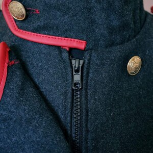 May include: A dark gray jacket with a red leather trim around the collar and a black zipper. The jacket has two gold buttons on the front.