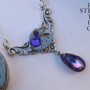 May include: A silver necklace with a steampunk-style pendant featuring a watch face with gears and a purple crystal. The pendant is attached to a silver chain with a teardrop-shaped purple crystal dangling from the bottom.
