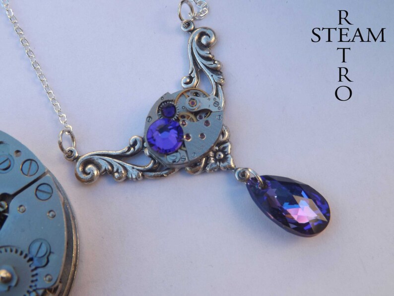 May include: A silver necklace with a steampunk-style pendant featuring a watch face with gears and a purple crystal. The pendant is attached to a silver chain with a teardrop-shaped purple crystal dangling from the bottom.
