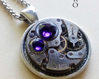 May include: Silver-toned pendant necklace featuring a circular pendant with watch gears and two purple gemstones. The pendant has the text "STEAM RETRO" on the top right. The necklace chain is also silver.