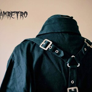May include: A black shirt with a dark green hue, featuring a silver buckle and straps on the back. The straps are in a Y-shape and have multiple silver buckles and rings.