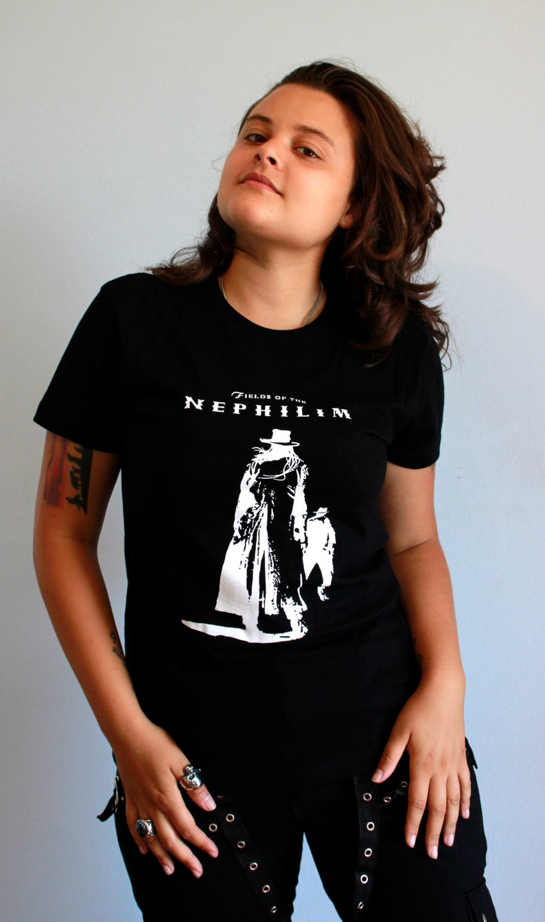 May include: A person wearing a black t-shirt with a white graphic of a figure in a top hat and the text "Fields of the Nephilim". The person is wearing black pants with silver grommets.