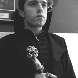 May include: A man in a black coat with a skull-shaped walking stick and rings on his fingers. The background is a white wall with a bat decoration.