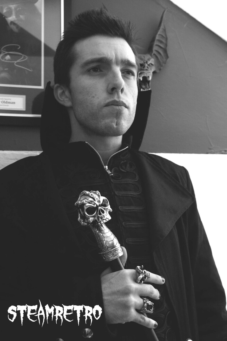 May include: A man in a black coat with a skull-shaped walking stick and rings on his fingers. The background is a white wall with a bat decoration.
