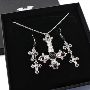 Puede incluir: Un conjunto de collar y pendientes de plata con un colgante de cruz invertida adornado con una rosa negra y detalles de cristal negro. Las joyas se presentan en una caja de terciopelo negro.