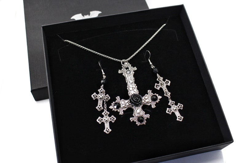 Puede incluir: Un conjunto de collar y pendientes de plata con un colgante de cruz invertida adornado con una rosa negra y detalles de cristal negro. Las joyas se presentan en una caja de terciopelo negro.