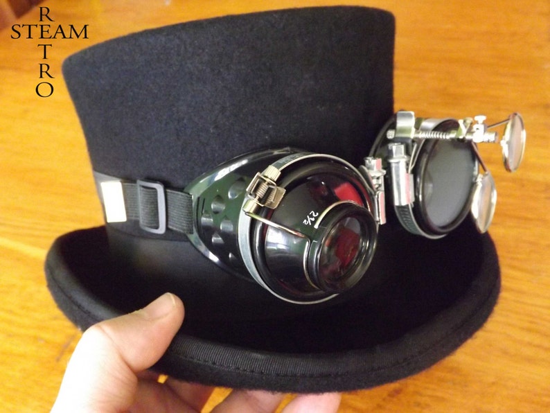 Peut inclure: Un chapeau haut de forme noir avec une paire de lunettes steampunk attach&eacute;es. Les lunettes ont une lentille grossissante d'un c&ocirc;t&eacute; et une lentille ronde de l'autre.