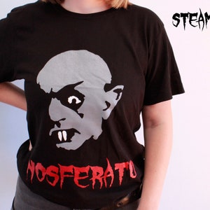 May include: Black t-shirt featuring a white graphic of a vampire's face and the text "NOSFERATU" in red.