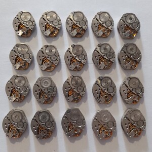 May include: A collection of 20 silver-toned watch movements with intricate gears and mechanisms. The movements are arranged in a grid pattern on a white surface.