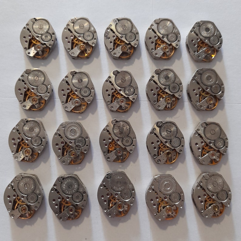 May include: A collection of 20 silver-toned watch movements with intricate gears and mechanisms. The movements are arranged in a grid pattern on a white surface.