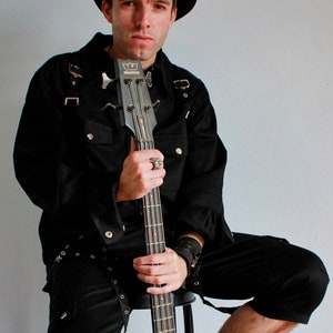 May include: A person wearing a black cowboy hat and a black shirt with a black bass guitar. The guitar has a black body and a silver headstock. The person is sitting on a stool and has a black belt with silver buckles. The text "STEAMRETRO" is in the upper right corner of the image.