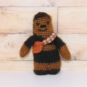 Op de afbeelding: Een gehaakt bruin en zwart Chewbacca-personage met een bruin en oranje bandolero. Het personage staat op een wit oppervlak.