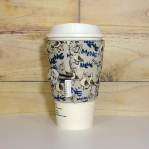 May include: A beige coffee cup cozy with a blue and white pattern of birds and the word "Mine" repeated. The cozy is on a white coffee cup.
