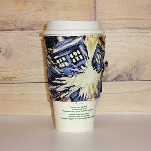 May include: A white coffee cup with a blue and yellow patterned sleeve featuring a stylized image of a spaceship. The sleeve has the text "Please use a cup sleeve. Contents may be hot." in English and French.