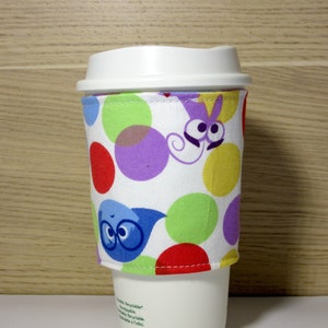 May include: A white coffee cup with a colourful fabric sleeve. The sleeve has a polka dot pattern with red, yellow, green, purple and blue dots. There are cartoon characters on the sleeve, including a blue character with glasses and a purple character with a swirl.