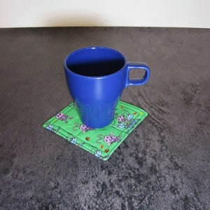 Może przedstawiać: Niebieski ceramiczny kubek stoi na zielonej, pikowanej podkładce z nadrukiem kresk&oacute;wek z mr&oacute;wkami. Podkładka ma białą obw&oacute;dkę.