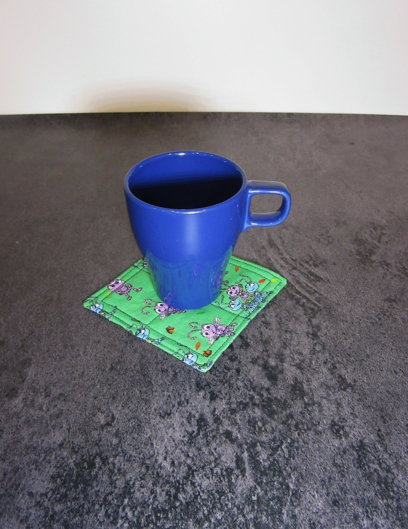 Może przedstawiać: Niebieski ceramiczny kubek stoi na zielonej, pikowanej podkładce z nadrukiem kresk&oacute;wek z mr&oacute;wkami. Podkładka ma białą obw&oacute;dkę.