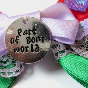 Op de afbeelding: Een zilveren bedel met de woorden "part of your world" erop gegraveerd. De bedel is bevestigd aan een paarse strik van satijnen lint met groene en rode strikken van satijnen lint en kant.