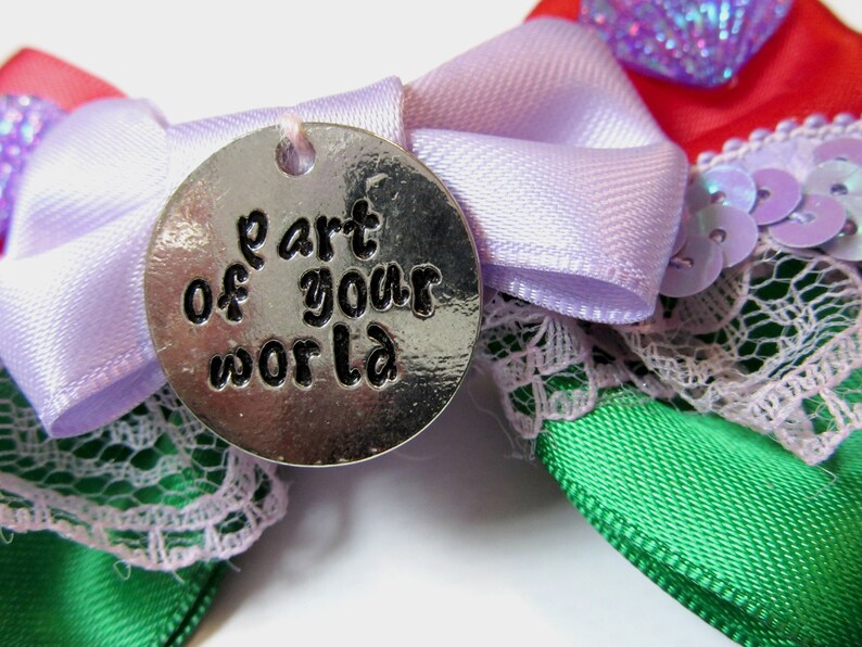 Op de afbeelding: Een zilveren bedel met de woorden "part of your world" erop gegraveerd. De bedel is bevestigd aan een paarse strik van satijnen lint met groene en rode strikken van satijnen lint en kant.