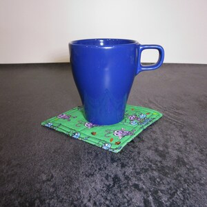 Może przedstawiać: Niebieski ceramiczny kubek stoi na zielonej, materiałowej podkładce z wzorem kresk&oacute;wkowych robak&oacute;w. Podkładka leży na ciemnoszarym tle.