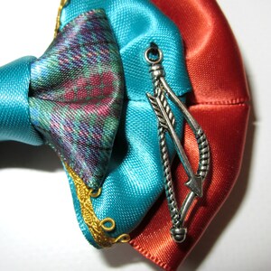 May include: A turquoise and orange satin bow with a silver arrow charm. The bow has gold trim and is tied with a knot.